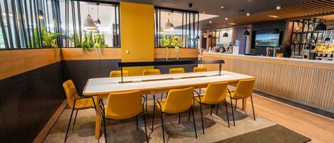 Una sala de conferencias moderna con una mesa larga y sillas amarillas, un área de bar con televisor y ventanas amplias con plantas.
