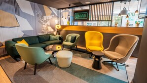 A modern lounge area with a green sofa, yellow and beige chairs, a small round table, and a 'ON AIR' sign. - Holiday Inn Bordeaux-Merignac by IHG (Merignac)