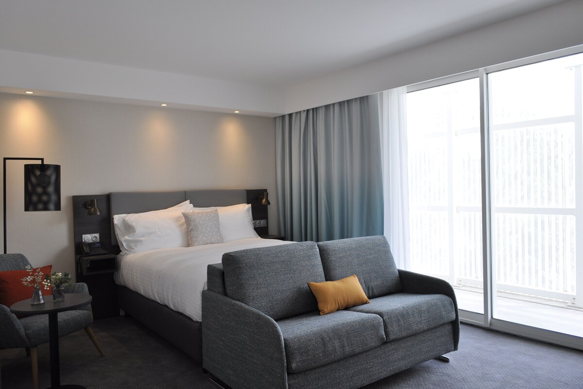 Ein modernes Hotelzimmer mit einem großen Bett, einem grauen Sofa und Blick durch ein großes Fenster nach draußen.