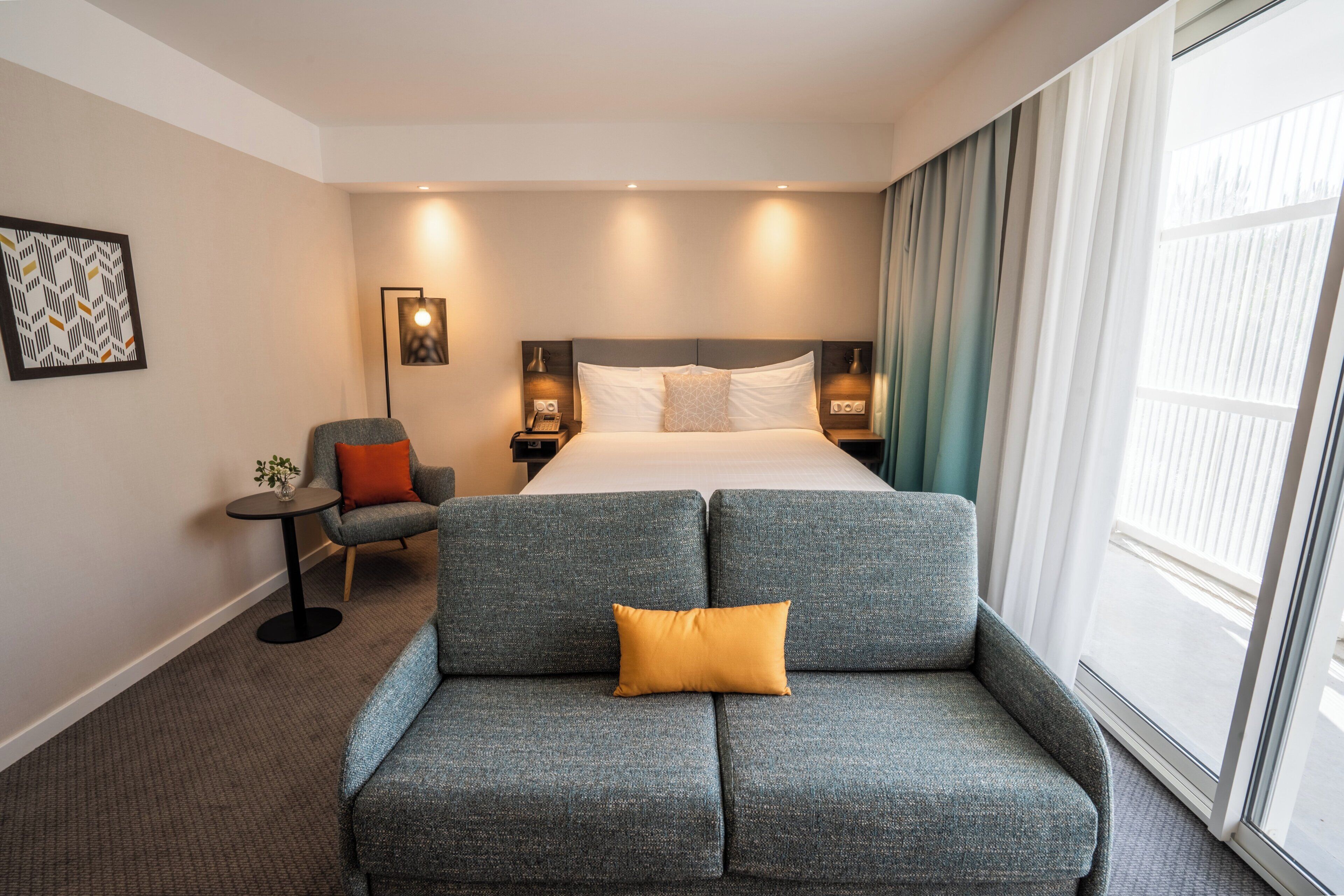 A hotel room with a bed, a grey sofa, a small table, and a chair.