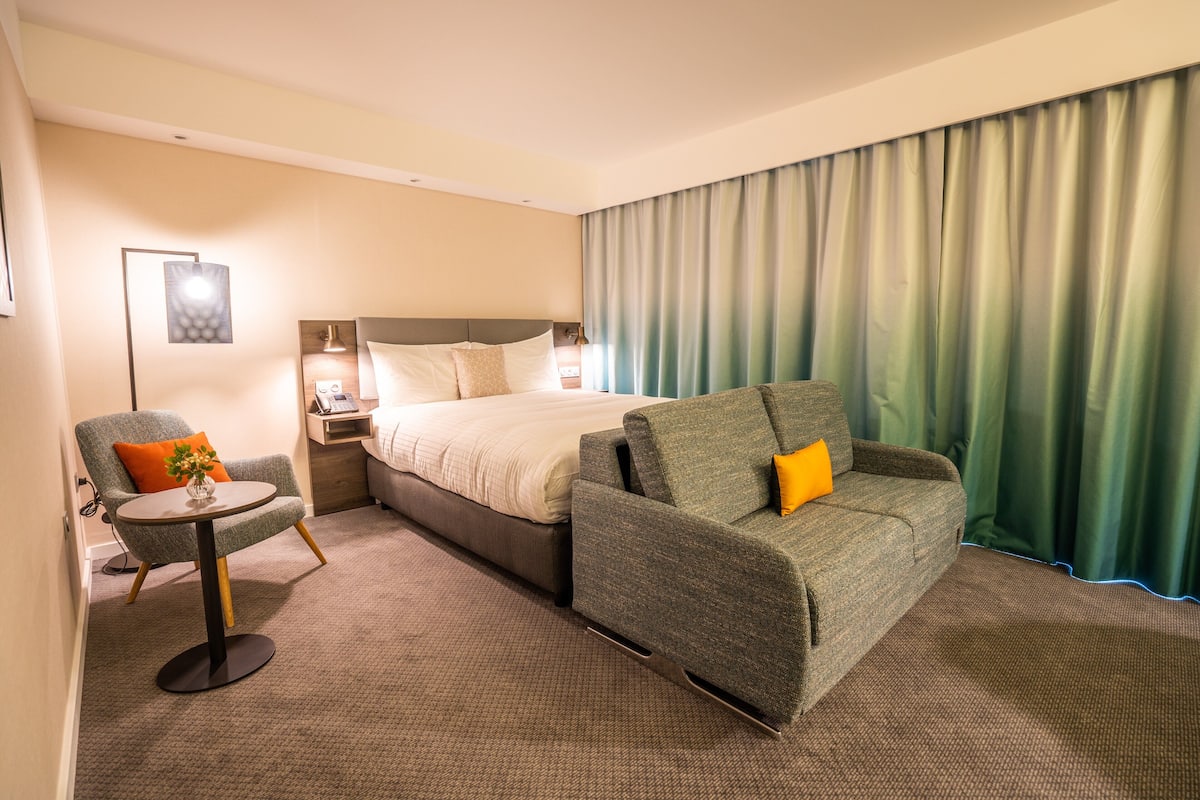 A hotel room with a bed, a sofa, a chair, a small table, and a large window with curtains.