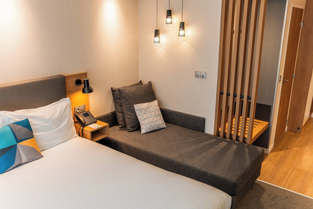 A hotel room with a large bed, a smaller sofa, a bedside table with a lamp, and a phone.