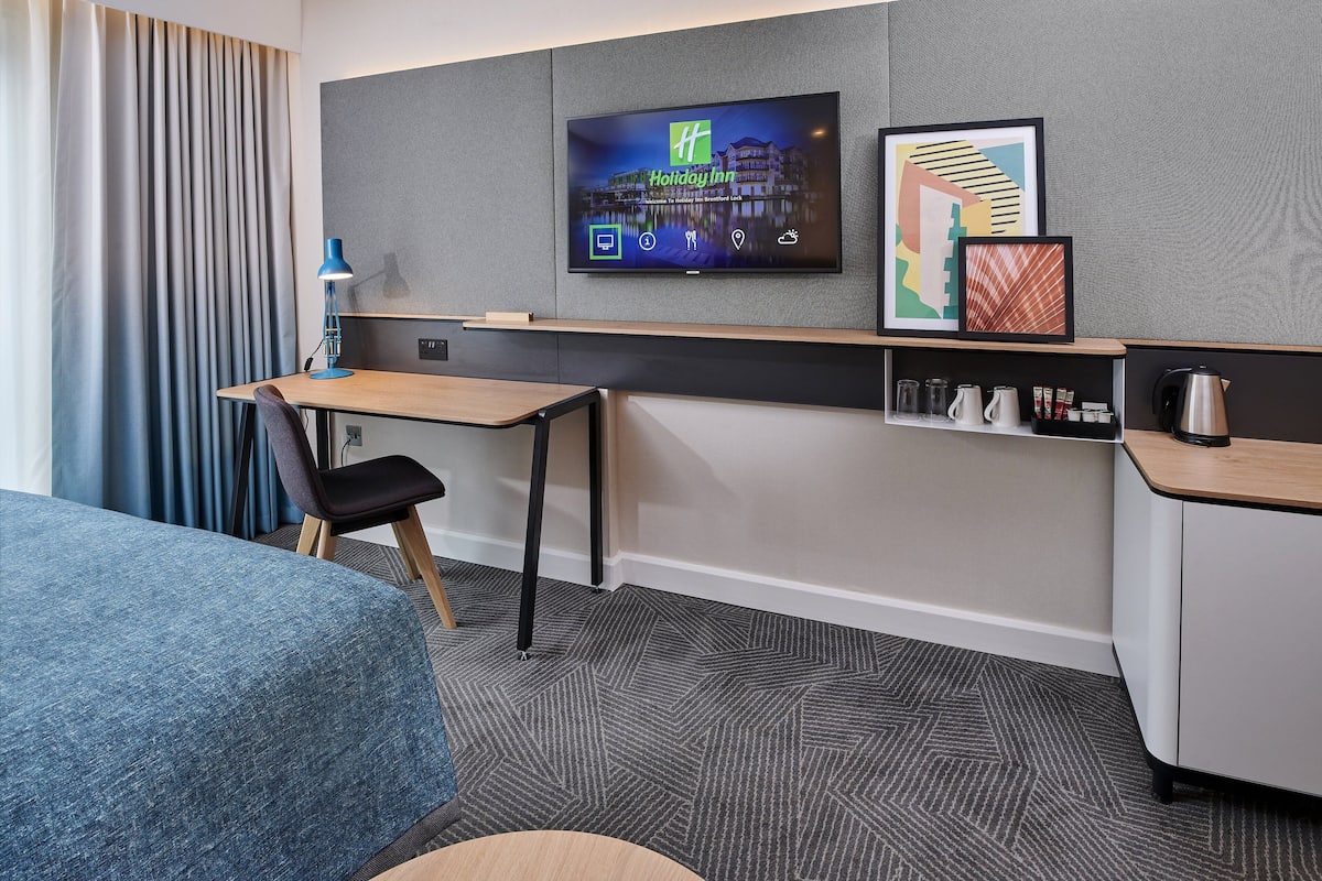 A hotel room with a bed, a desk, a chair, a TV mounted on the wall, and a coffee machine.