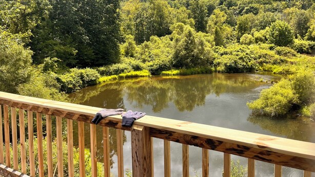 Une terrasse en bois surplombe une rivière paisible, avec une végétation luxuriante et des collines en arrière-plan.