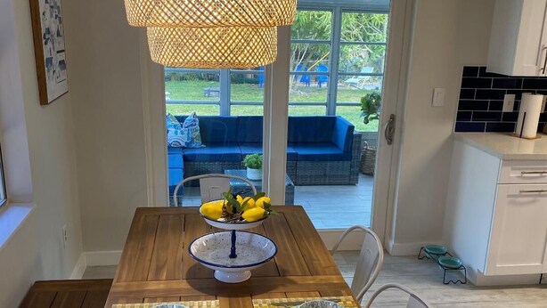 Un coin repas avec une table en bois dressée pour deux personnes, un luminaire suspendu et une vue sur un espace extérieur aménagé avec des sièges bleus.