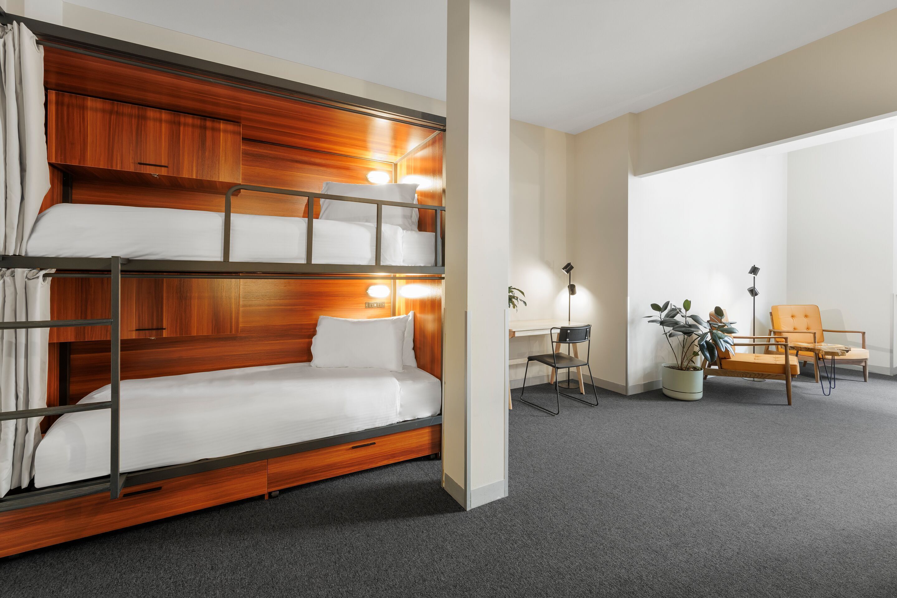 A bunk bed with wooden headboard and bedside lamps, a desk with a chair, and a potted plant in a hotel room.
