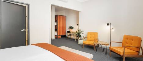 Habitación de hotel con cama, una manta naranja, una silla de madera, una mesita y una planta en maceta.