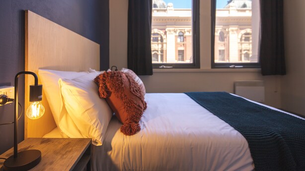 A hotel room with a bed, a nightstand with a lamp, and a window with a view of a building. - Bounce Melbourne (Melbourne)