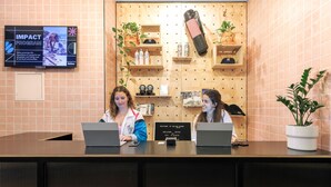 A reception desk with two staff members, a wall-mounted TV displaying an 'IMPACT PROGRAM' ad, wooden shelves with plants and bottles, and a sign welcoming guests. - Bounce Melbourne (Melbourne)