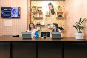 Una recepción con dos empleados, un televisor empotrado mostrando un anuncio del 'IMPACT PROGRAM', estanterías de madera con plantas y botellas, y un cartel dando la bienvenida a los huéspedes.