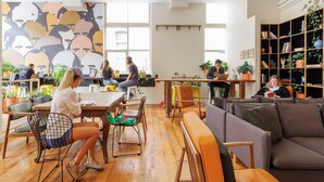 A modern co-working space with wooden floors, a variety of seating options including tables, chairs, and a couch, and a large bookshelf filled with books and plants. - Bounce Melbourne (Melbourne)