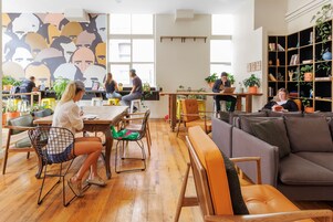 Un espacio de coworking moderno con pisos de madera, diversas opciones de asiento como mesas, sillas y un sofá, y una amplia estantería llena de libros y plantas.