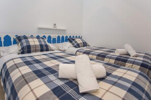A hotel room with two beds, each with a checkered bedspread and pillows. There is a white shelf above the beds and a white towel rolled on each bed.