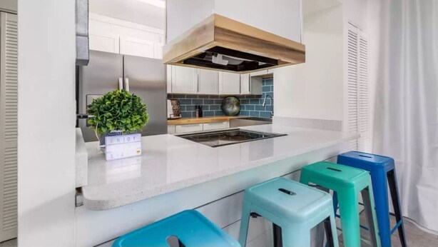 A modern kitchen with white cabinets, a stainless steel range, a blue tiled backsplash, and a wooden hood. There are three colorful stools and a potted plant on the counter. - The Beach Bungalow - In the Heart of Destin, FL!   (Destin)