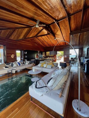 A spacious living area with a large white sectional sofa, a glass coffee table, and a pool. The room features wooden ceilings and a modern kitchen in the background.