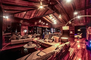 A spacious, well-lit lounge area with a high ceiling and wooden paneling. There is a seating area with sofas and a coffee table, and a bar area with a counter and stools.