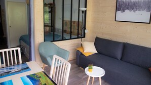 Um quarto aconchegante com um sofá cinzento, uma poltrona azul, uma pequena mesa branca com uma planta, e uma grande janela com vista para árvores.