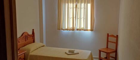 A small, cozy room with two single beds, a wooden chair, and a window with curtains.