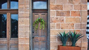 Una puerta de madera con un adorno de flores, una maceta grande con plantas y un muro de ladrillo.