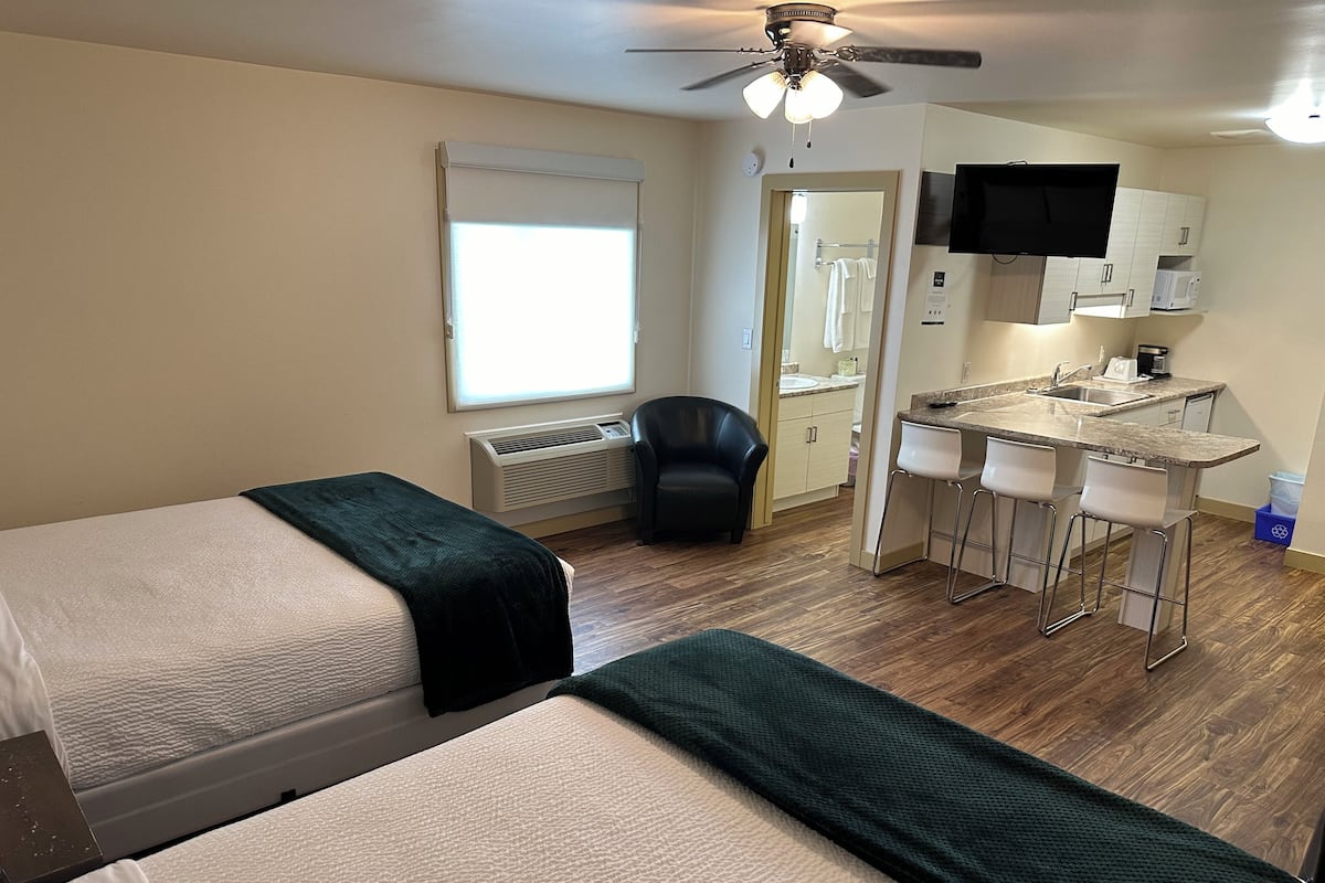A hotel room with two beds, a desk with a chair, a ceiling fan, a window with blinds, and a kitchenette.