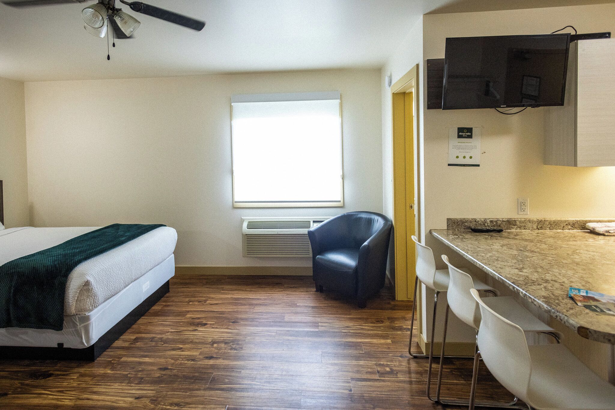 Habitación de hotel compacta con cama, un pequeño escritorio con silla, una ventana con persianas, ventilador de techo y televisor de pantalla plana montado en la pared.