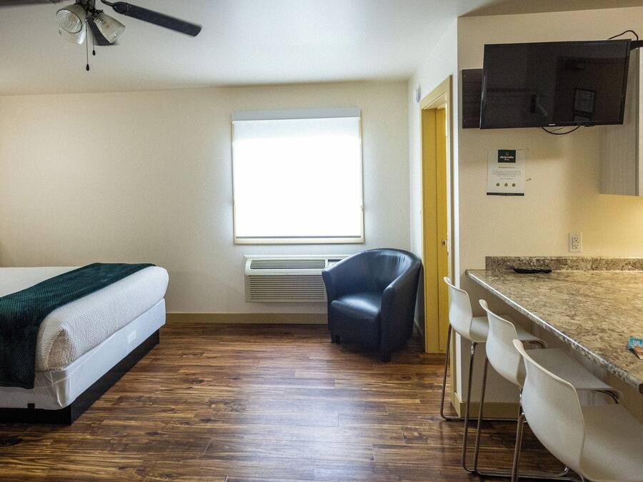 Habitación de hotel compacta con cama, un pequeño escritorio con silla, una ventana con persianas, ventilador de techo y televisor de pantalla plana montado en la pared.