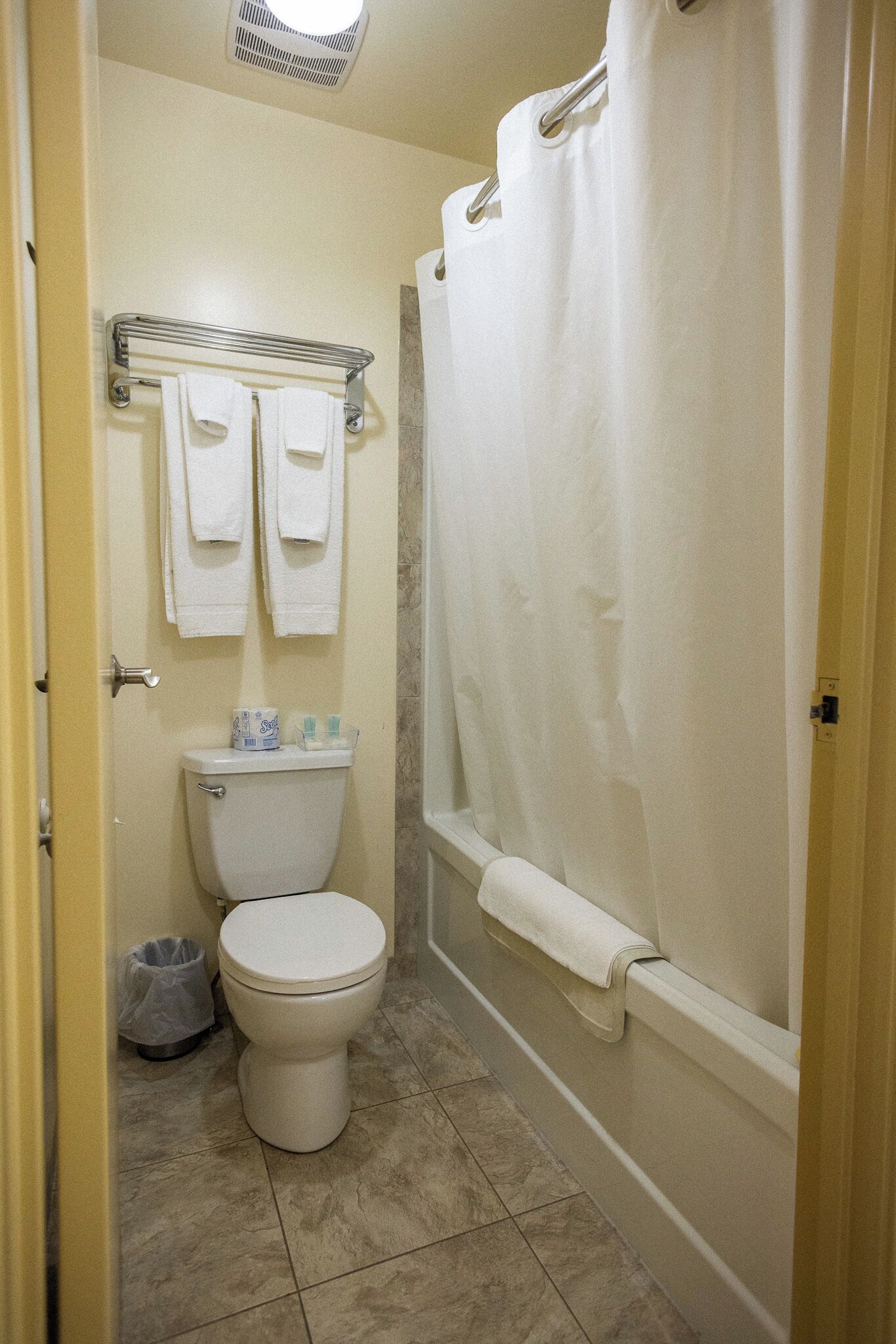 A bathroom with a toilet, a towel rack with towels, a shower curtain, and a trash can.
