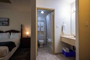 Ein Hotelzimmer mit Bett, Nachttisch mit Lampe, Bad mit Toilette, Dusche, Waschbecken und einem blauen Mülleimer.