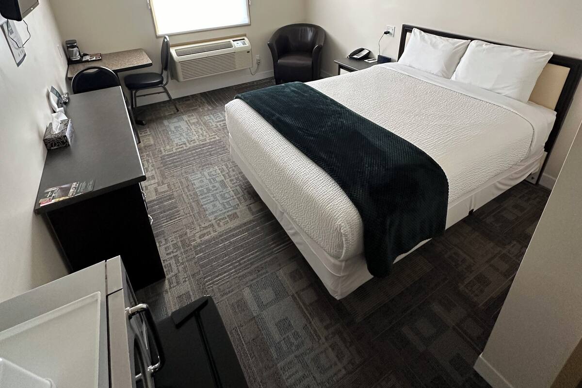 A hotel room with a bed, a desk, a chair, a small table, a heater, a window, and a wall-mounted phone.