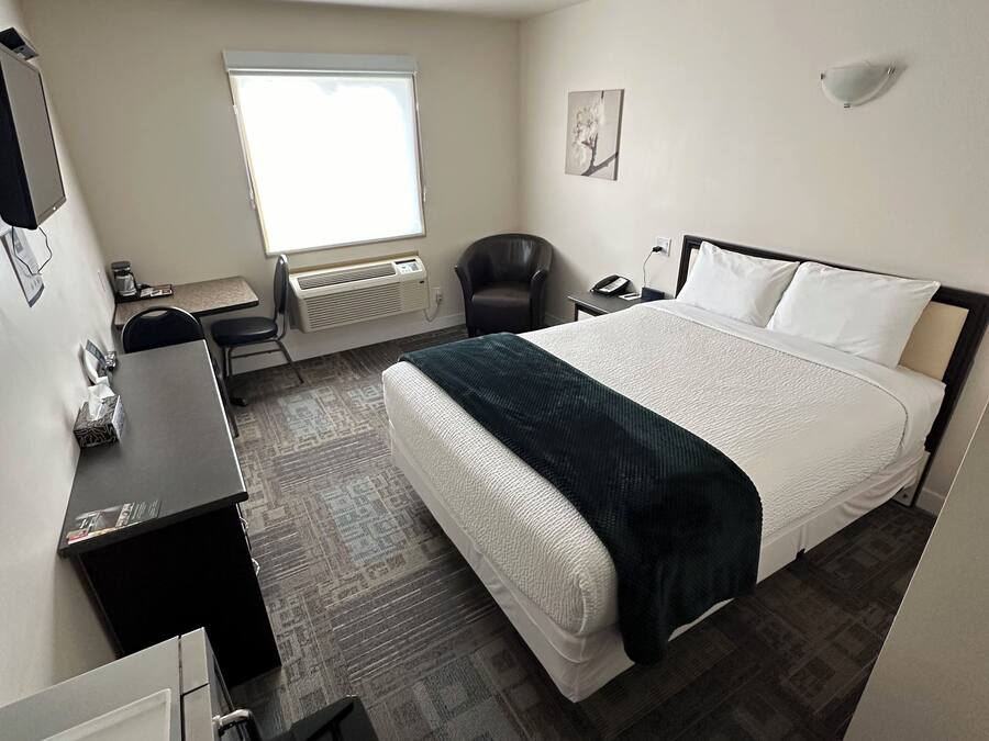 A hotel room with a bed, a desk, a chair, a small table, a heater, a window, and a wall-mounted phone.