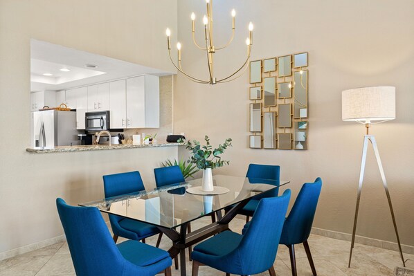 A modern dining area with a glass table, blue chairs, a chandelier, a decorative mirror, and a floor lamp.