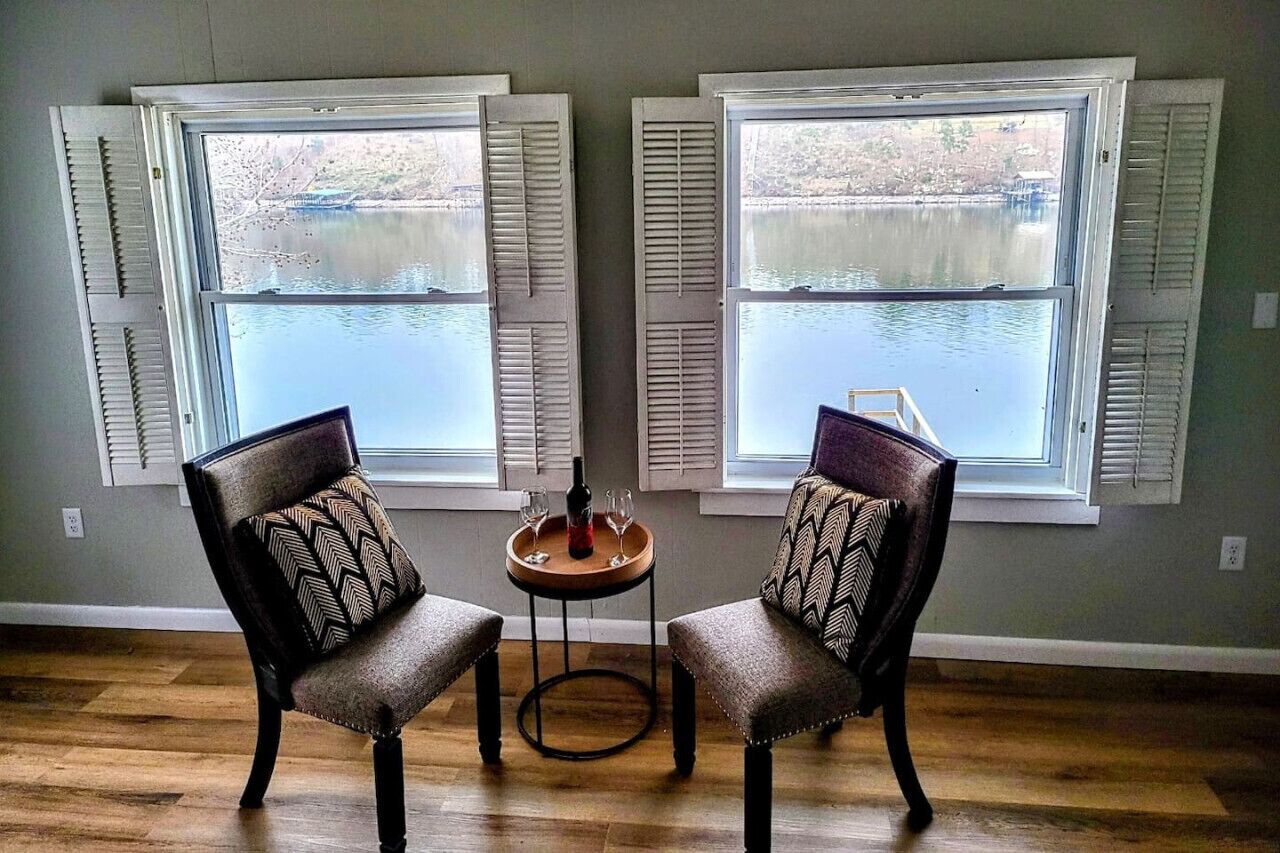 Una habitación con dos sillas frente a una mesa con una botella y vasos, ventanas amplias con contraventanas y vista a un cuerpo de agua.