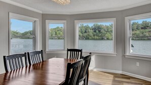 Un comedor con una gran mesa de madera y sillas, tres ventanas amplias con vista al lago y un candelabro.