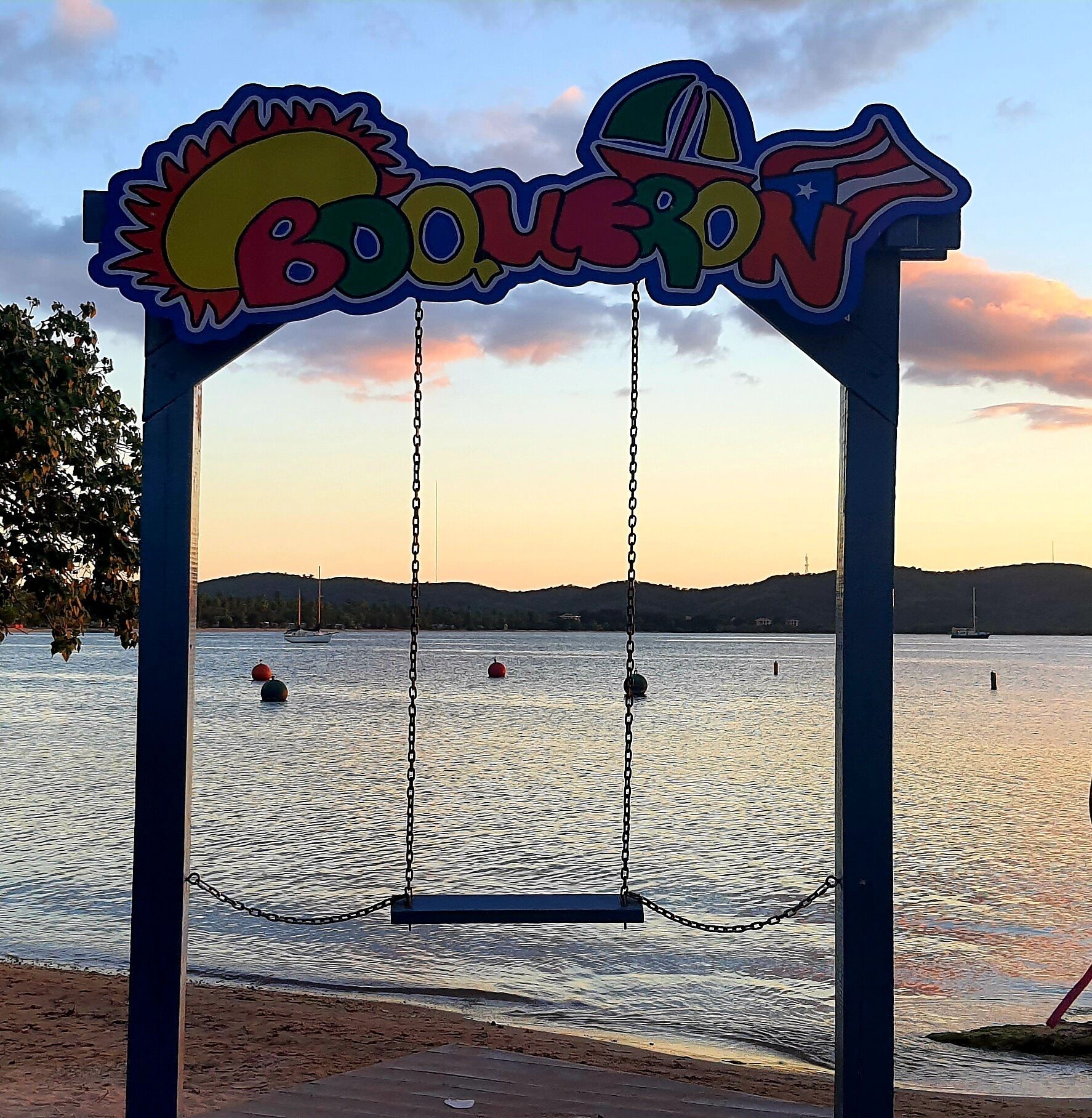 A swing set with a sign that reads "BOULEVON" against a backdrop of a calm lake and distant hills during sunset.