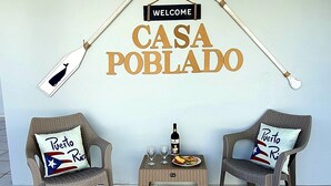 Una pared con un cartel de bienvenida y las palabras 'CASA POBLADO' en letras grandes. Hay dos sillones de mimbre con cojines que llevan el diseño de la bandera de Puerto Rico. Una pequeña mesa con una botella y vasos se encuentra entre las sillas.