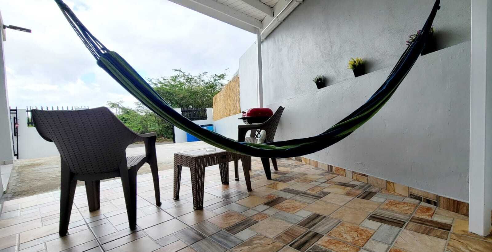 Un'amaca sospesa tra due sedie su un patio con pavimento piastrellato, un tavolino e una fioriera a parete.