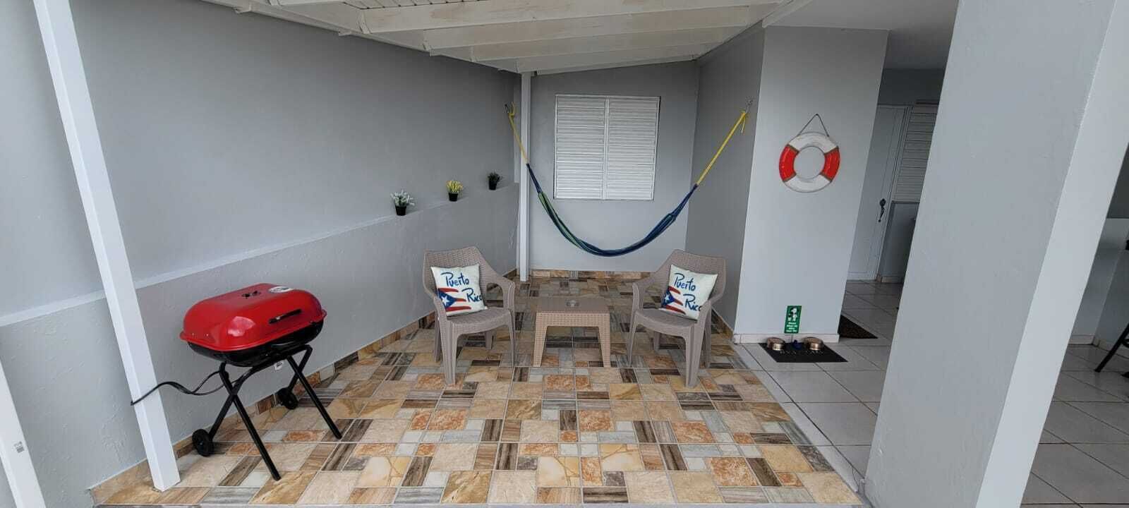 Un petit patio abrité, doté d’un sol carrelé, de deux fauteuils en osier avec des coussins, d’un barbecue rouge et d’un hamac.