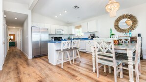 Une cuisine moderne avec des meubles blancs, un réfrigérateur en acier inoxydable, des tabourets de bar en bois et une table à manger avec des chaises.