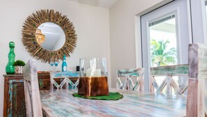 Une table à manger rustique, agrémentée d’un chandelier en cuivre, est entourée de chaises d’époque. Un miroir à cadre en bois est accroché au mur, et une fenêtre offre une vue sur des palmiers.