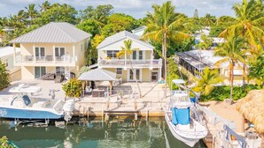 Une propriété située au bord de l’eau, avec des maisons à deux étages, un quai où sont amarrés des bateaux et une structure surmontée d’un toit de chaume.