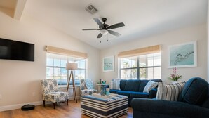Un salon avec un canapé bleu, un pouf à rayures, un fauteuil à motifs et un ventilateur de plafond.