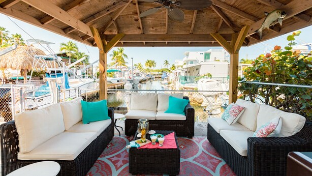 Un espace extérieur abrité, aménagé avec des meubles en osier, un ventilateur de plafond et offrant une vue sur les bateaux et les bâtiments en arrière-plan.