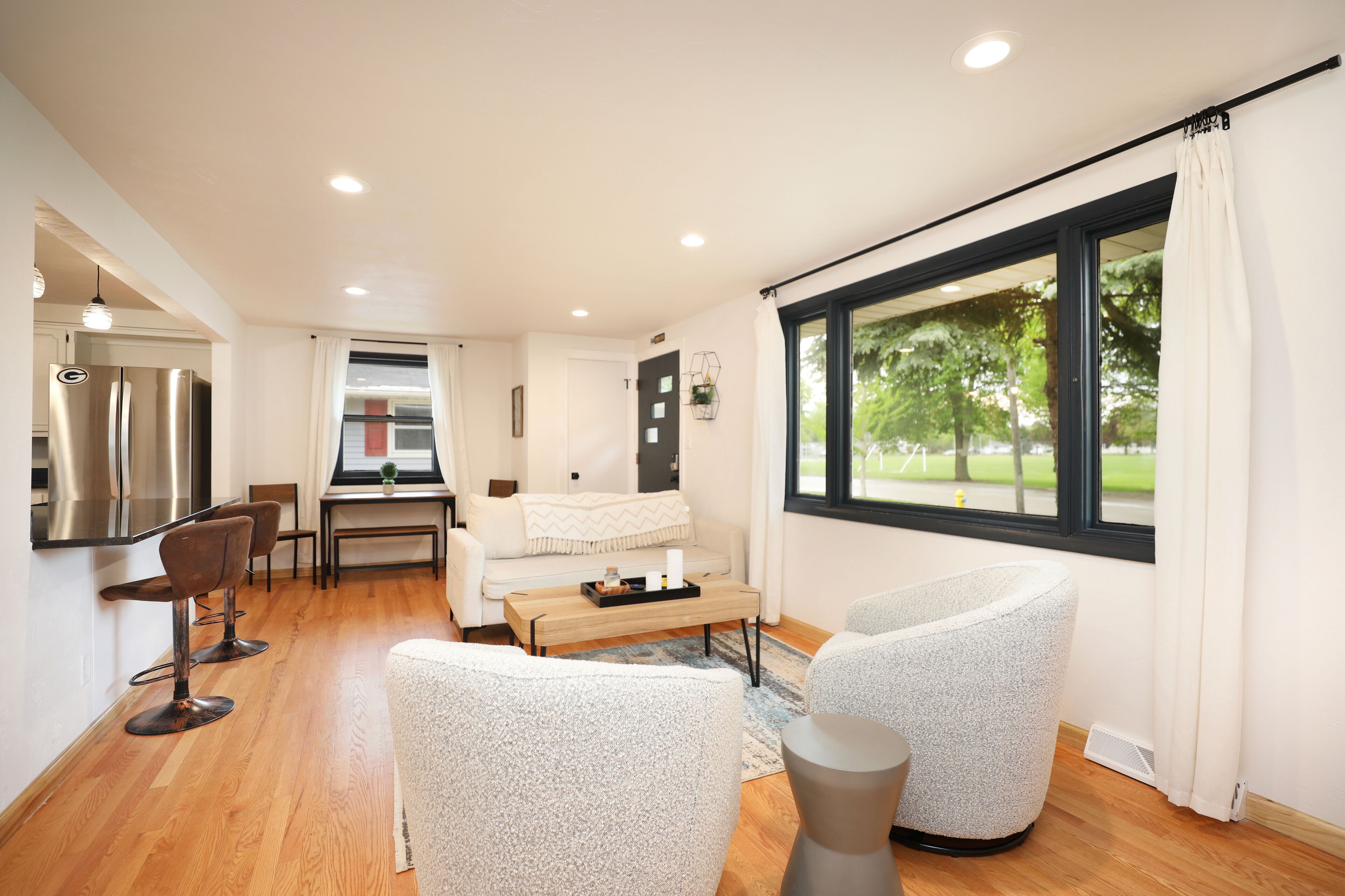 A modern kitchen with stainless steel appliances, a dining area with a wooden table and chairs, a cozy seating area with a sofa and armchairs, and a large window with a view of greenery.