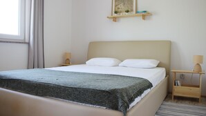 A neatly made bed with a grey blanket, a wooden bedside table with a lamp, and a small shelf with decorative items. - Secluded vacation house surrounded by nature (Žminj)