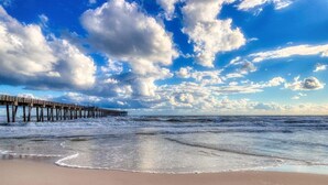 Una playa con un muelle de madera que se adentra en el océano, bajo un cielo parcialmente nublado.