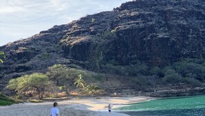 Ein Sandstrand mit Fußabdrücken, die zum Wasser führen, eine Person, die entlang geht, und eine sanfte Hügelkulisse mit vereinzelter Vegetation.