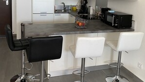 A modern kitchen with white cabinets, a central island with bar stools, a microwave, and a kettle. - Cosy apartment rental (SANTA LUCIA DI MORIANI)