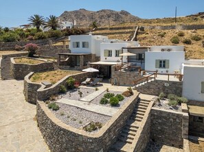 A white villa with a stone courtyard, outdoor seating, and a mountainous backdrop.