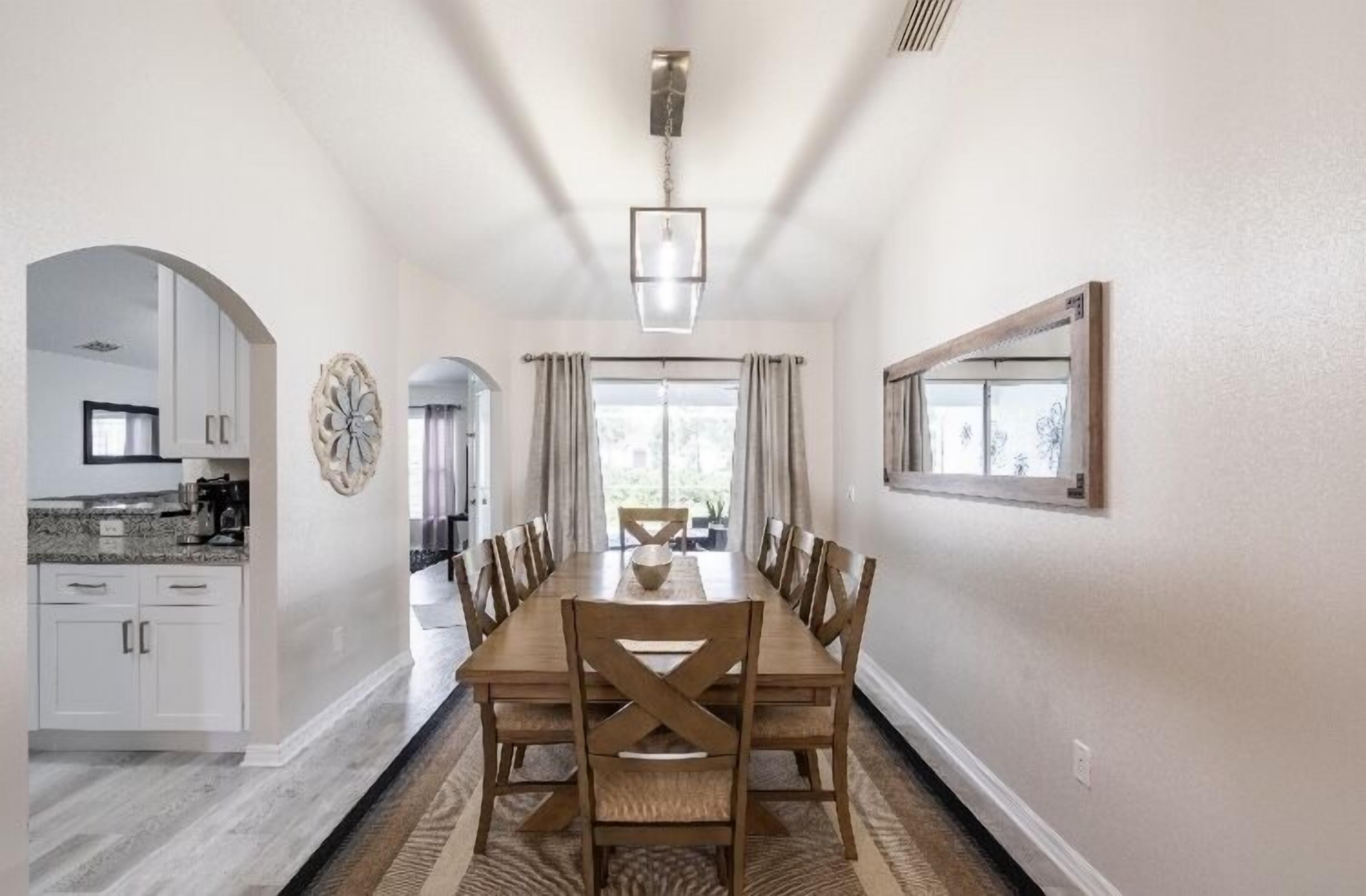 Une salle à manger avec une longue table, des chaises et un lustre. On peut apercevoir une cuisine sur la gauche, avec des placards et un plan de travail.