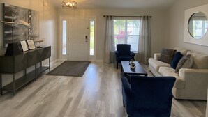 Un salon spacieux comprenant un canapé, deux fauteuils, une table basse et un téléviseur. Une porte donne accès à un balcon d’où l’on peut admirer la verdure.